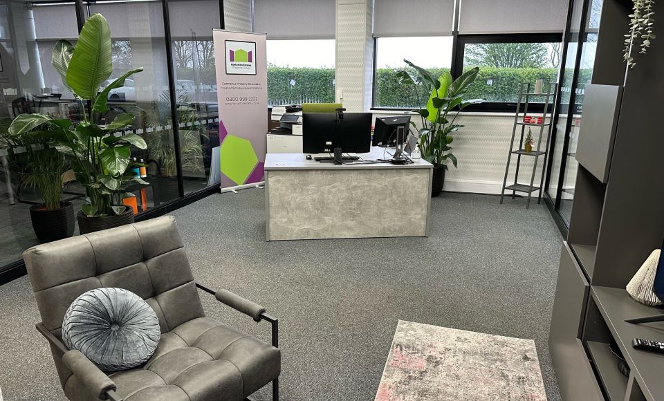 Modern office with grey carpet, a cushioned armchair, a desk with dual monitors, large potted plants, and a banner for an estate agency in the background.