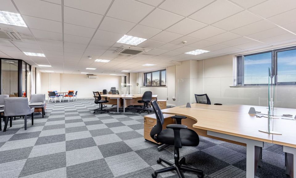 Spacious modern office with open-plan desks, black chairs, grey checkered carpet, and large windows providing natural light.