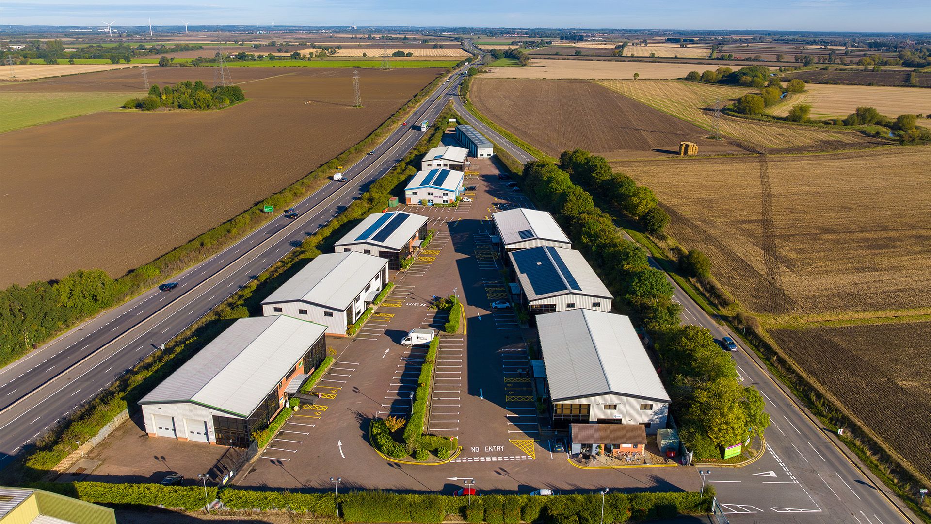 Aerial view of an industrial estate with multiple warehouse units, parking spaces, and a nearby highway surrounded by fields.