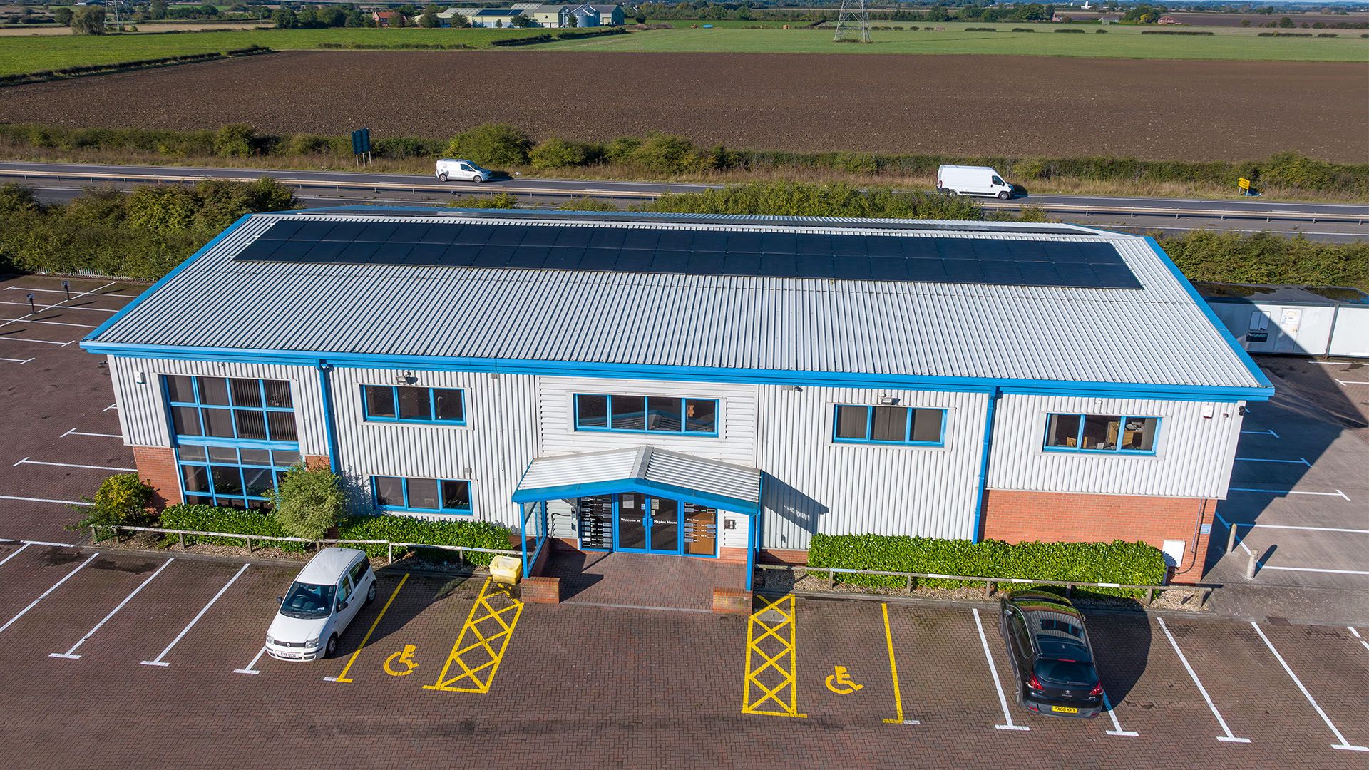 Aerial view of Mayden House, a modern office building with blue trim, surrounded by an empty car park and fields in the background.